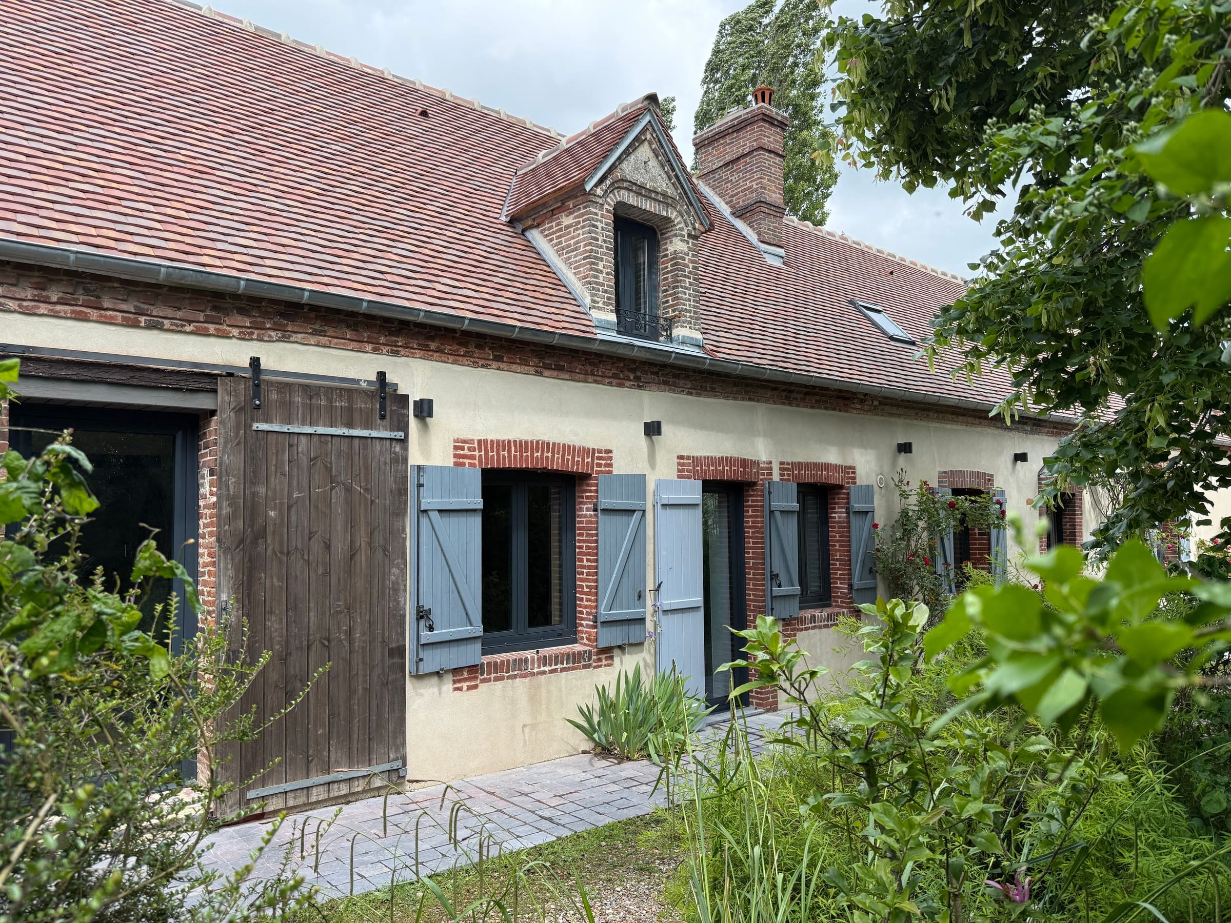 La façade sud en plein jour : toiture neuve, lucarne, porte de grange et volets bleus